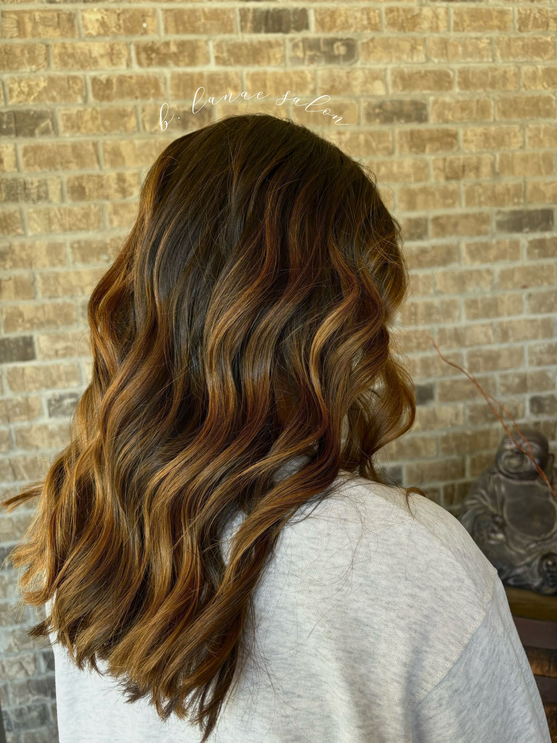 Woman with brown, wavy hair against a brick wall. Highlights are golden. She wears a light sweater.