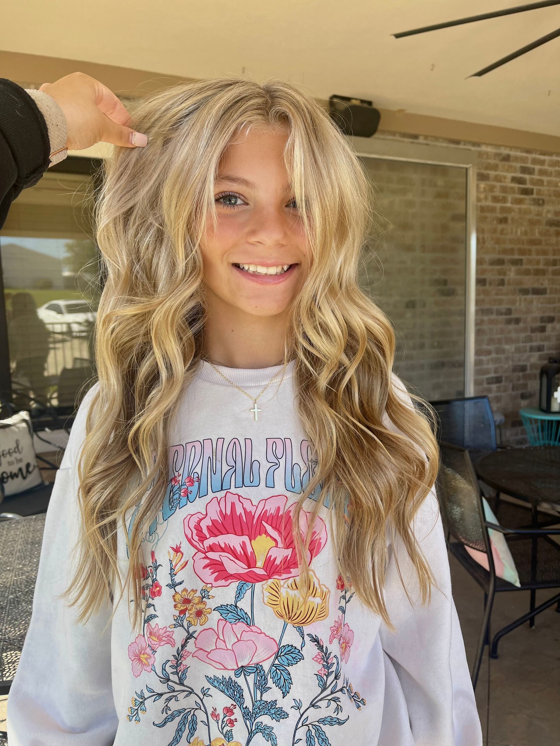 Blonde-haired teen smiles, wearing a floral sweatshirt, outdoors under a covered patio.