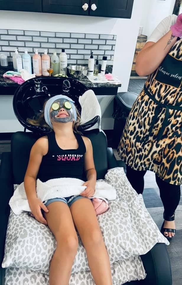 Girl with face mask at salon, sitting in chair, getting hair wash. Leopard-print apron and pillow.