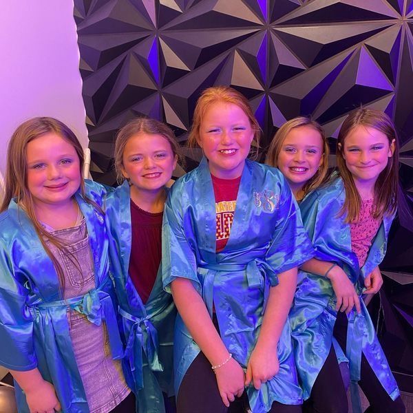 Five girls in blue robes, posing with makeup, in front of a geometric wall.