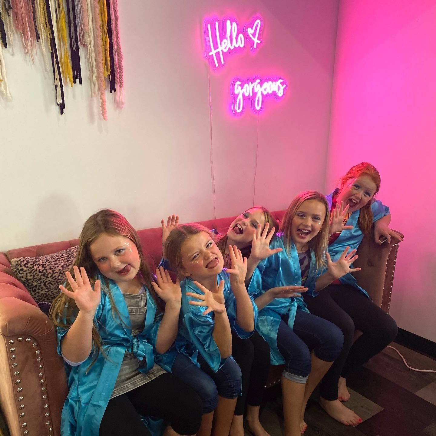 Five young girls in blue robes waving on a pink couch under a neon sign that says 