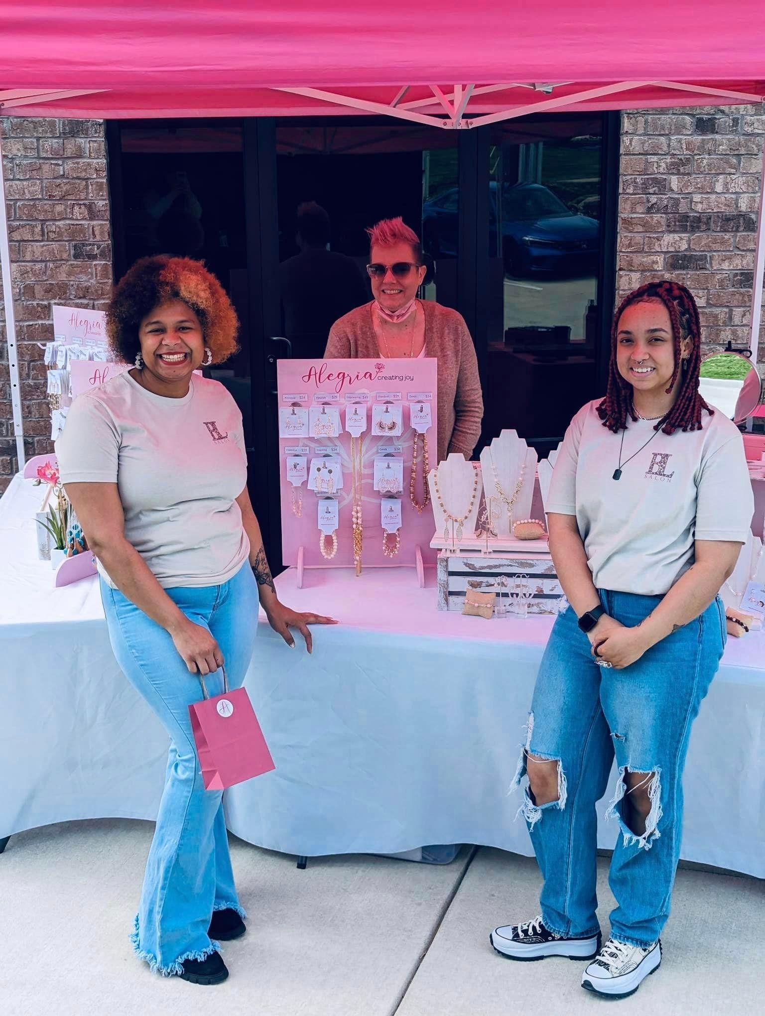 Three women at a jewelry booth under a pink tent. Tan shirts, jeans, jewelry display. Outdoor setting.