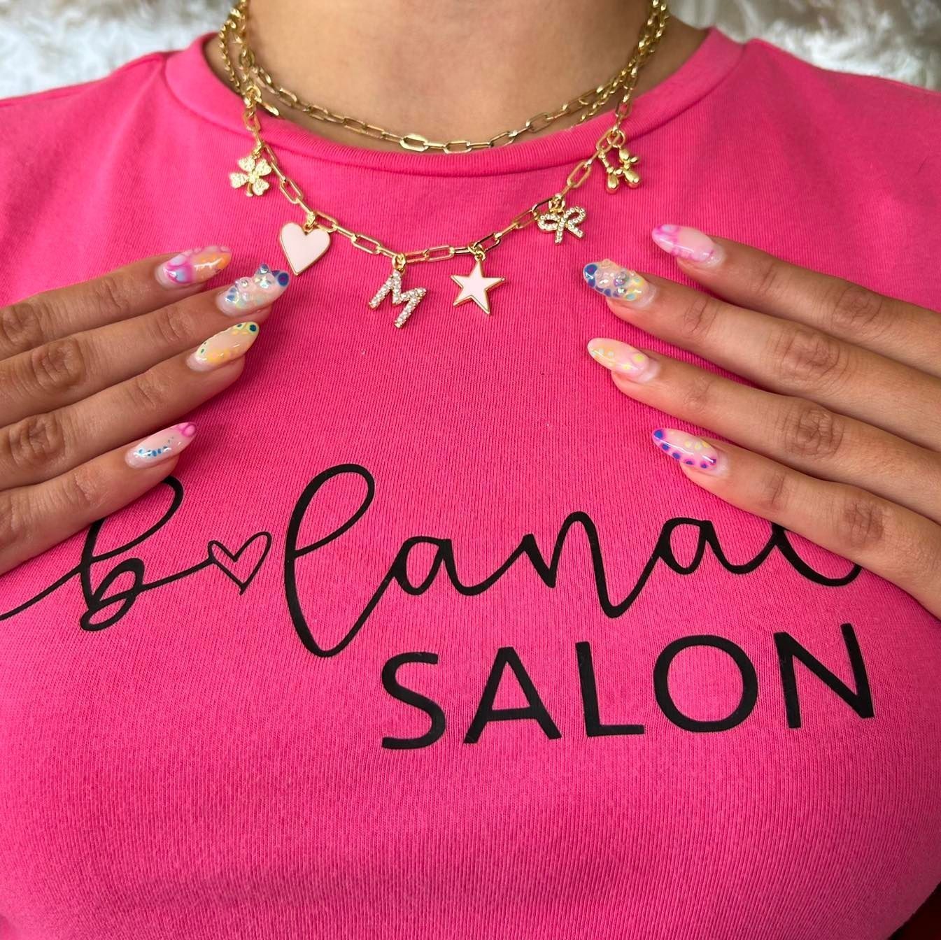 Person wearing a pink shirt and gold charm necklace. Nails are painted with pastel colors. Shirt reads 