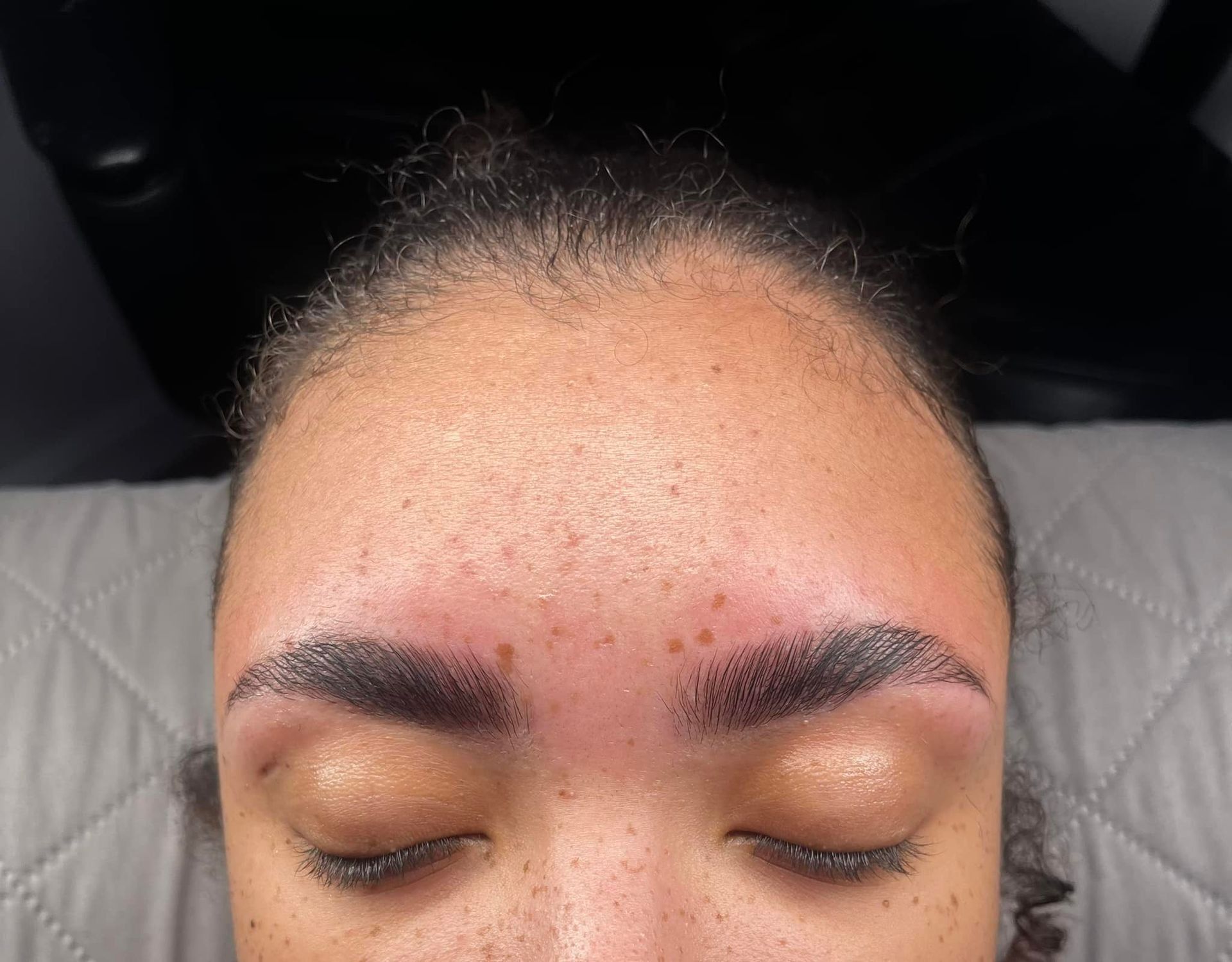 Person with closed eyes, freshly done dark eyebrow tattoos, light skin with slight redness.