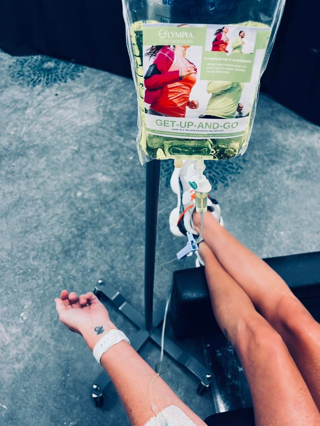 Person receiving IV fluids, arm and legs visible. IV bag hangs above, visible tattoo on wrist.