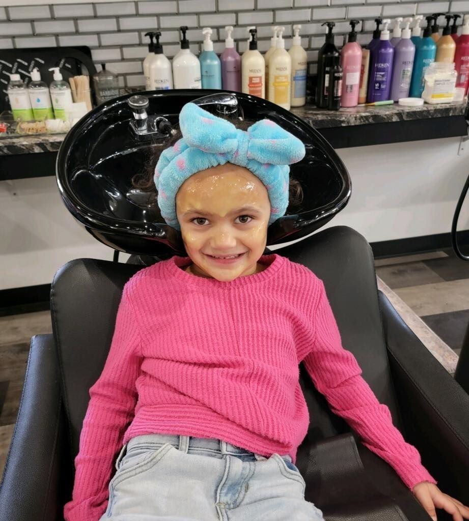 Girl in a salon chair with a face mask and headband, smiling at the camera.