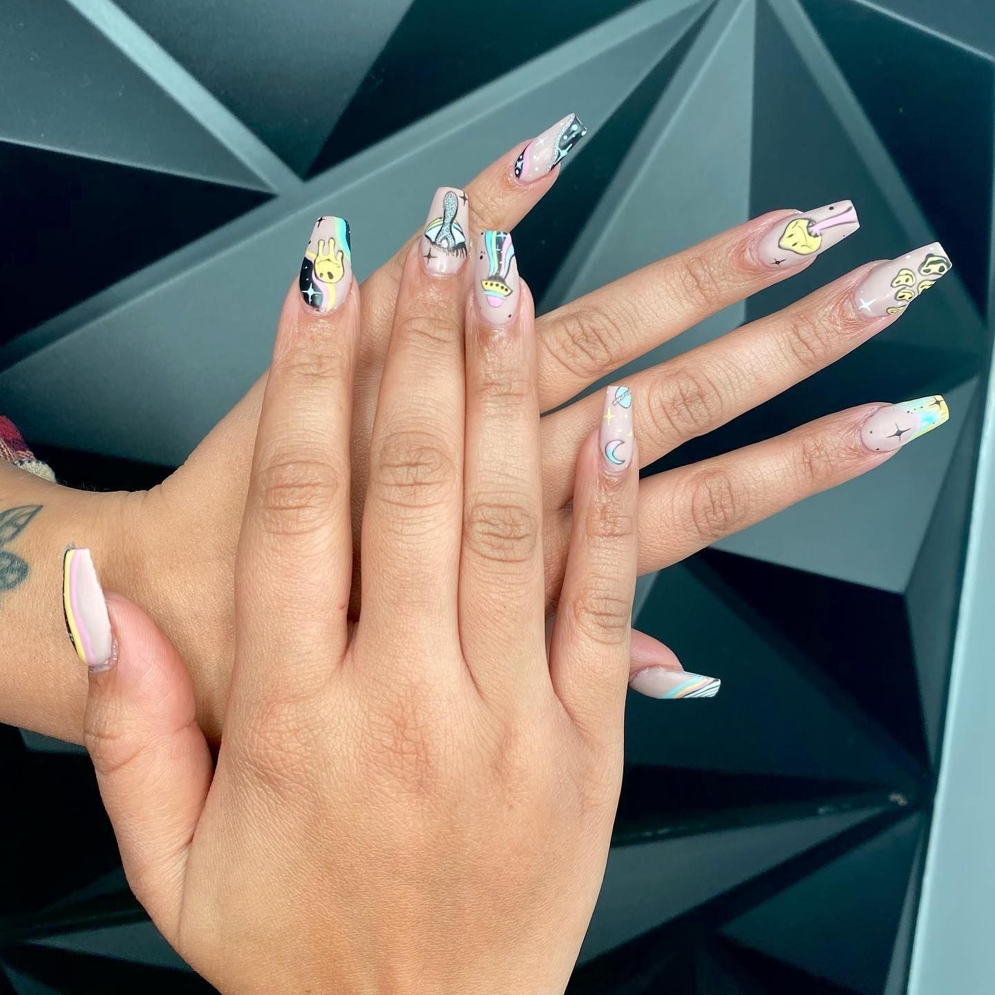 Hands with long, decorated acrylic nails. Each nail has a unique design with colorful art and gems.