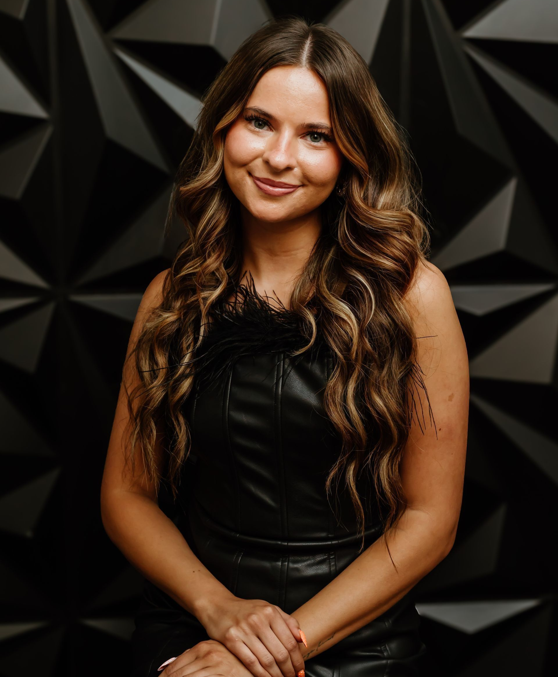 Woman with long wavy brown hair wearing a black leather dress smiles, posing against a geometric black wall.