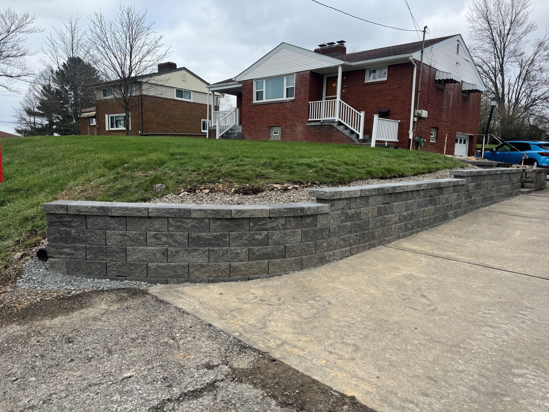 A gray, stacked-stone retaining wall sits along the edge of a concrete driveway in front of a brick house.