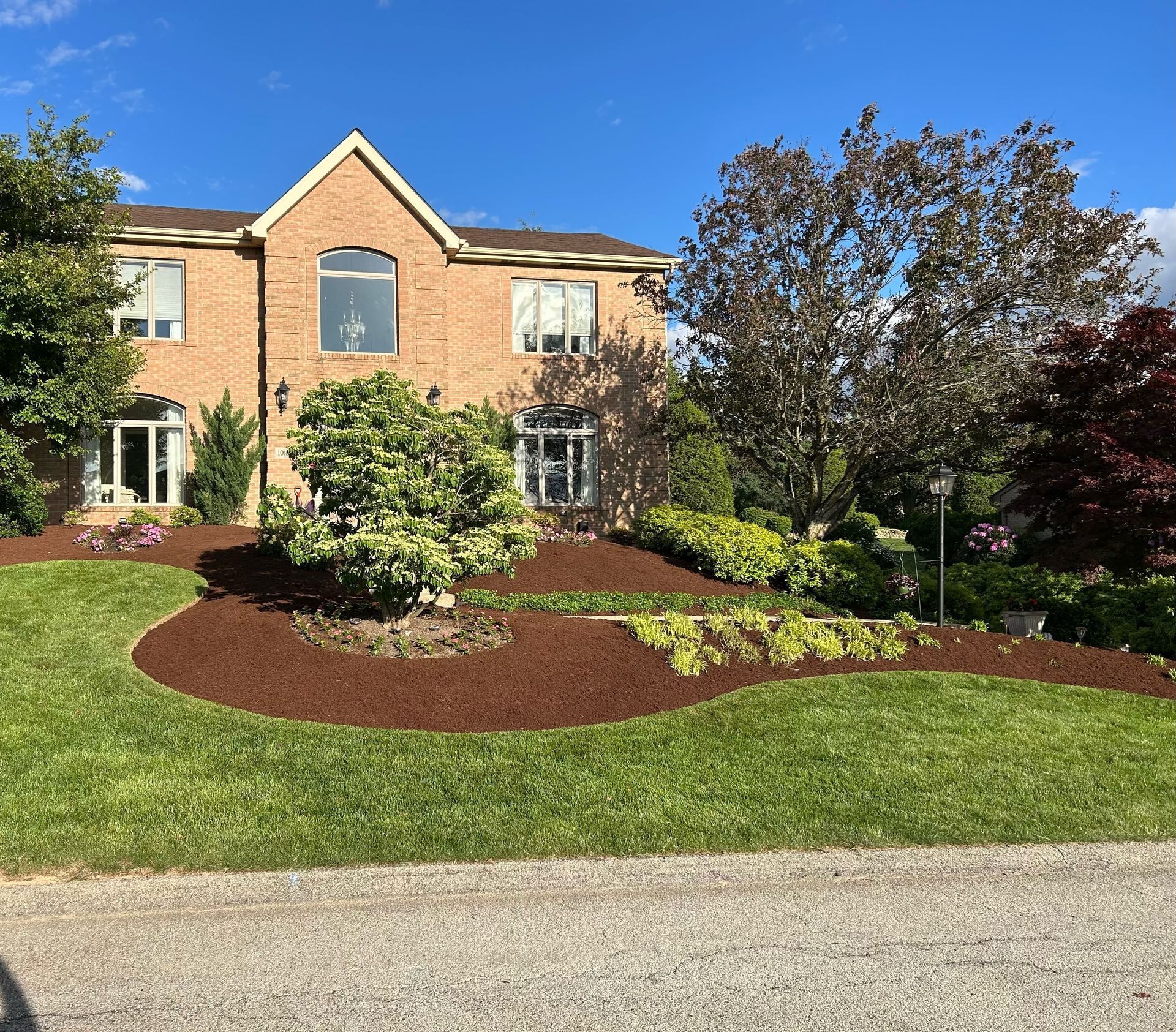Two-story brick house with landscaped front yard, mulch beds, green lawn, and blue sky.