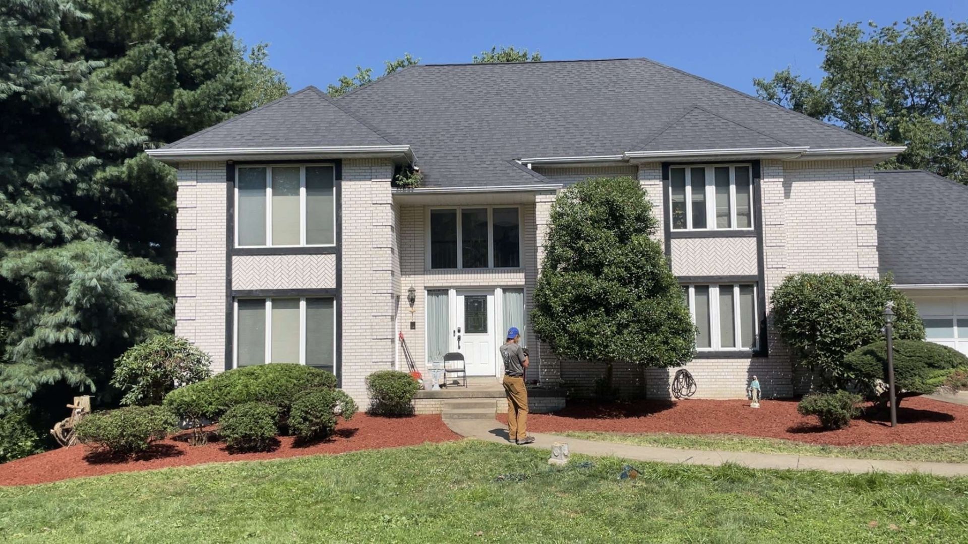 Two-story house with gray roof, white exterior, and a person standing in front, on a sunny day.