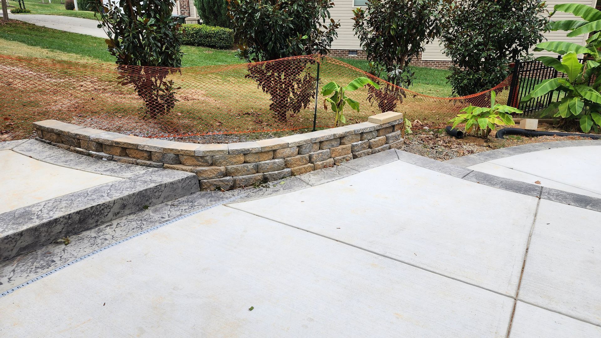 Curved brick retaining wall bordering concrete driveway with green bushes and lawn.