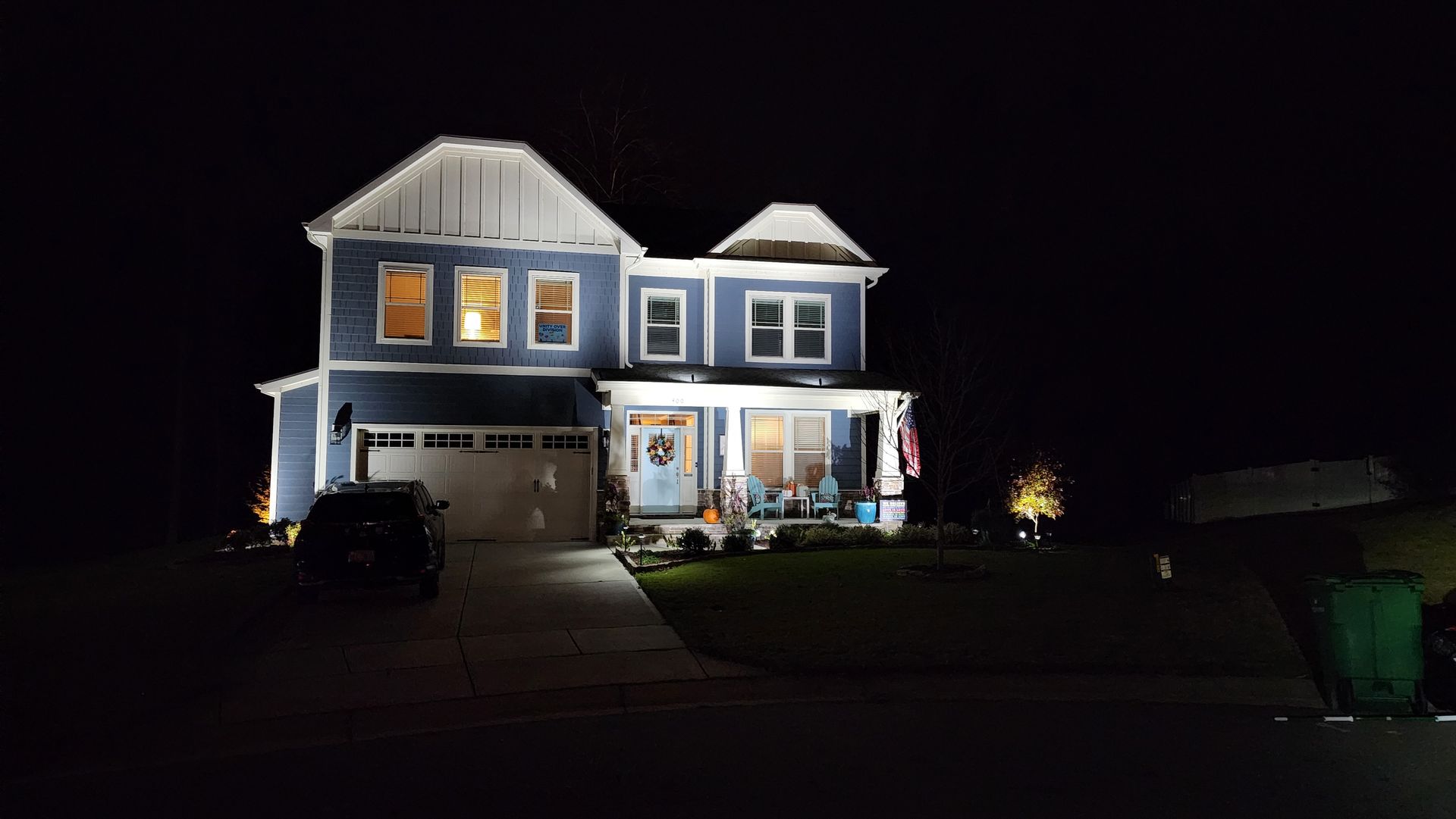 Two-story blue house at night, lit from inside with a car parked in front and a green trash can to the side.