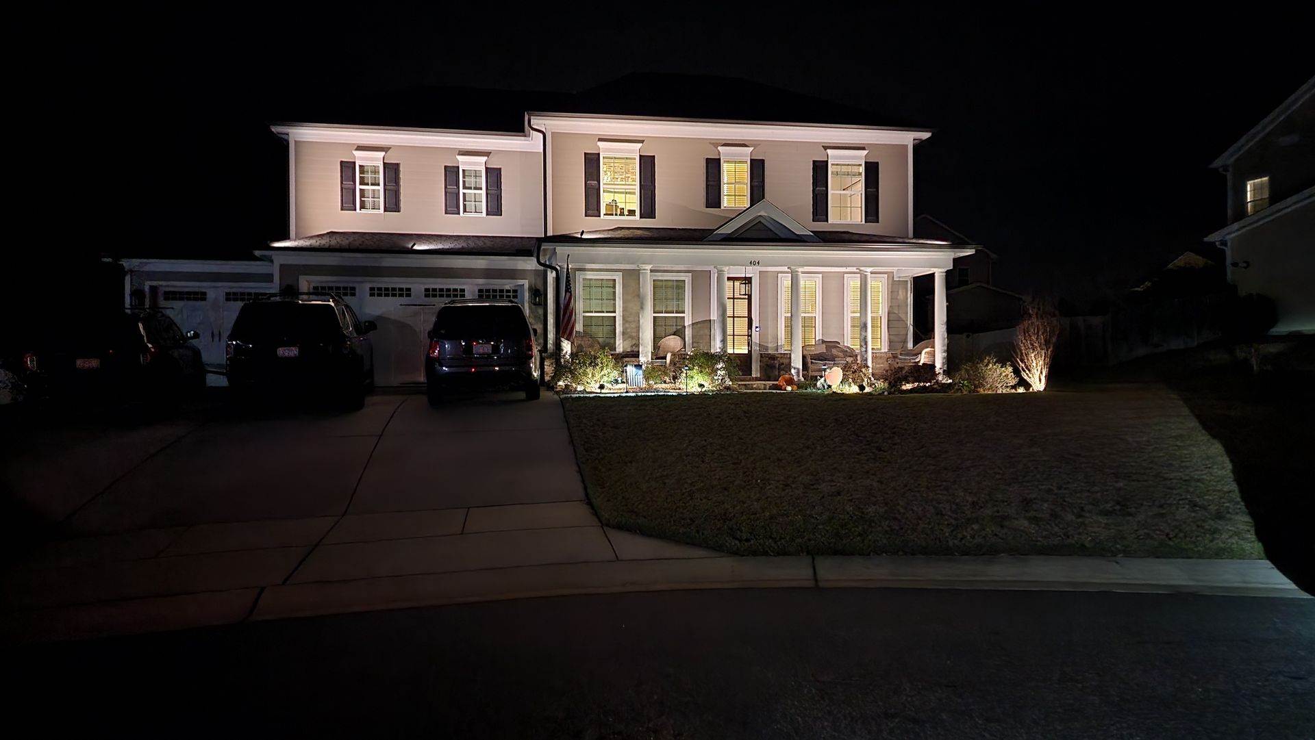A two-story house at night with lights on, illuminated driveway, cars parked, and lawn.