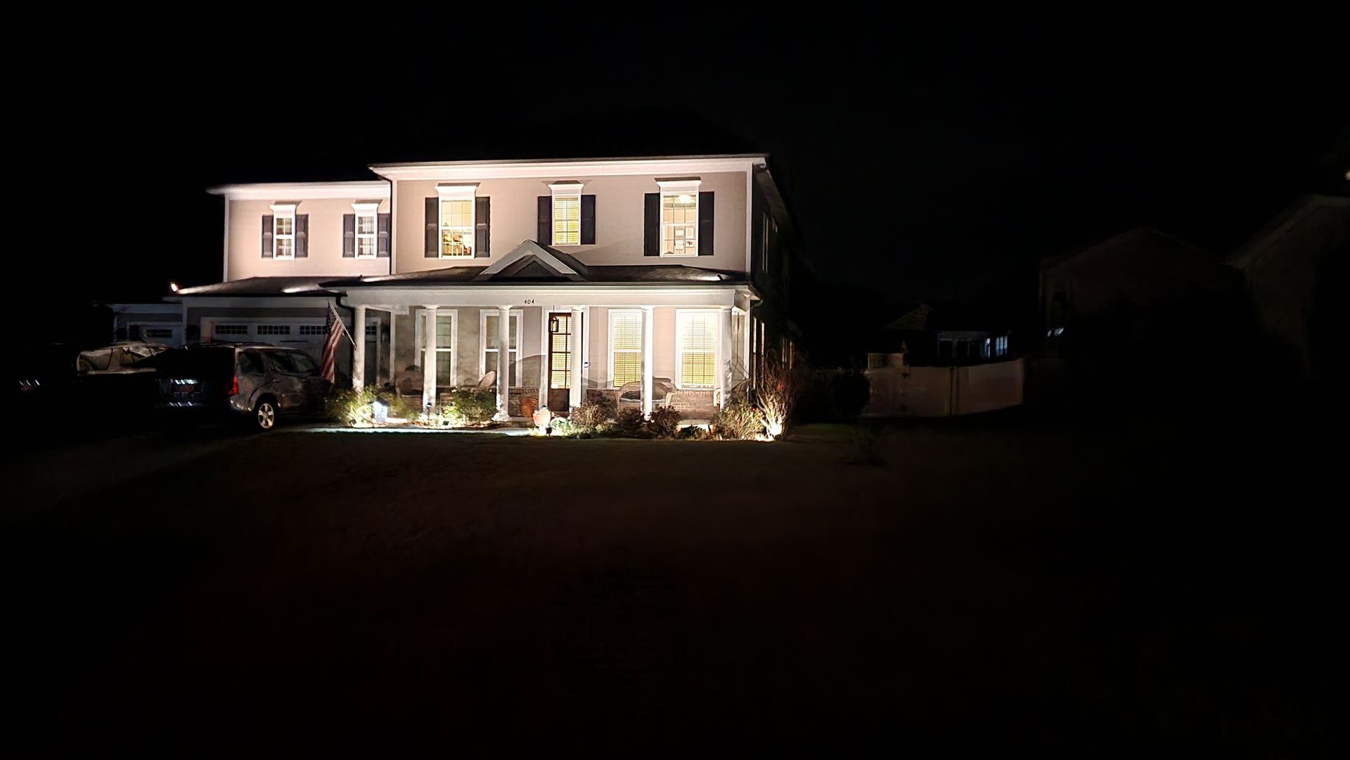 Two-story house at night with lights illuminating the porch and windows. Dark surroundings.