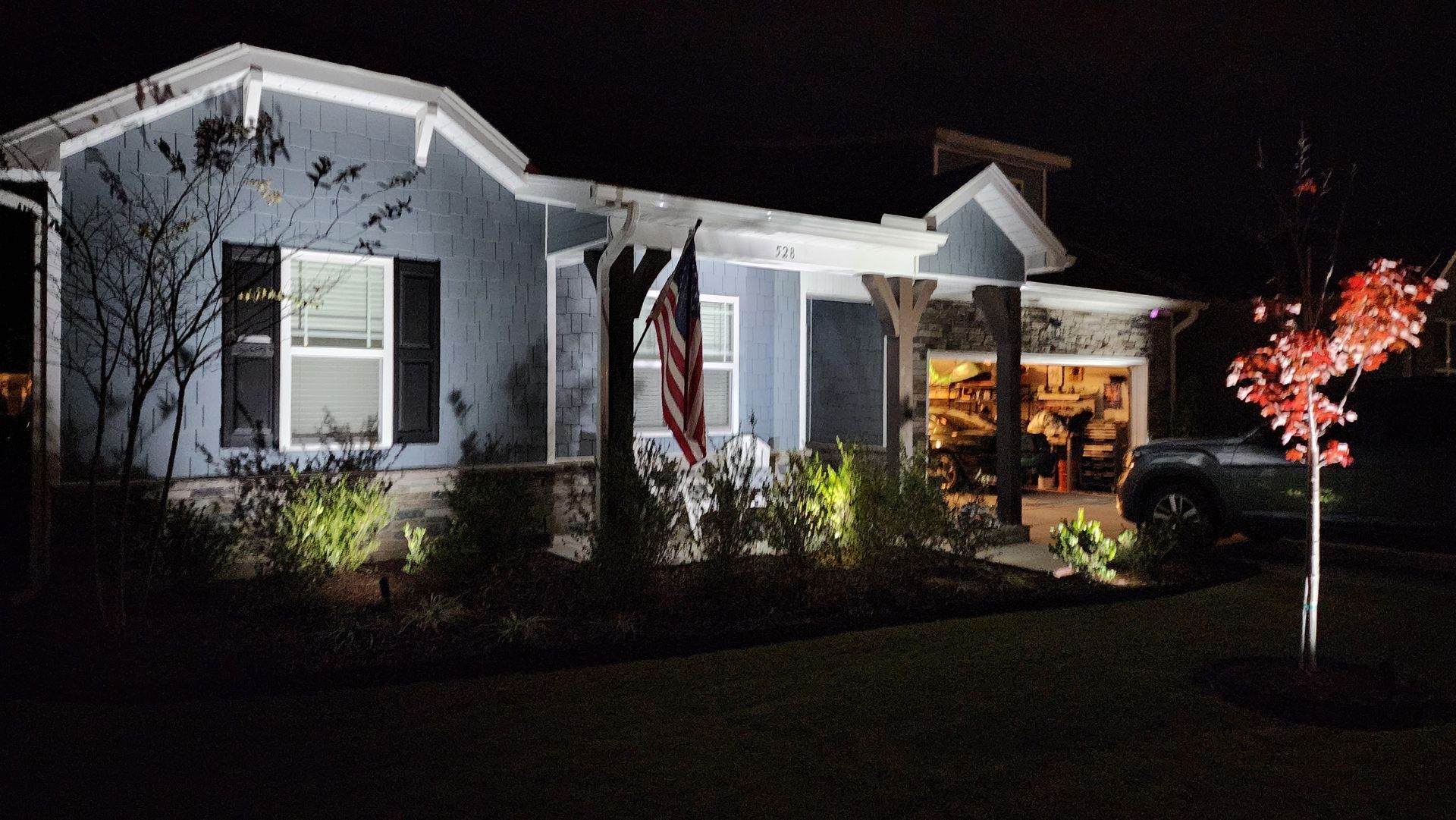Blue house at night with white trim, lit landscaping, and an American flag.