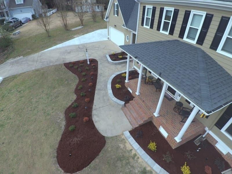 Tan house with porch and black shutters, brown mulch beds along driveway.