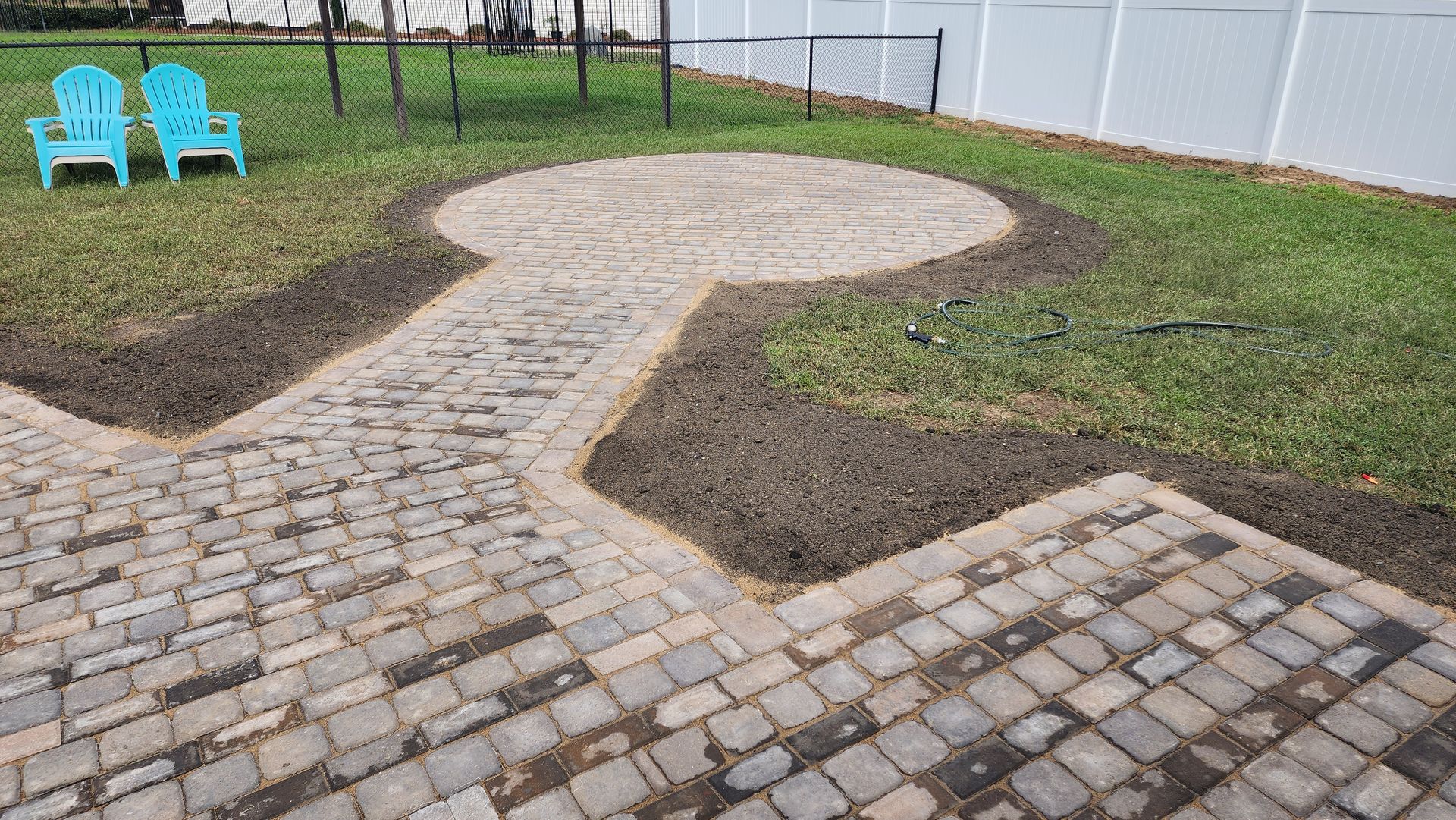 Paver patio with a circular area and pathway, flanked by grass. Two blue chairs sit in the yard.
