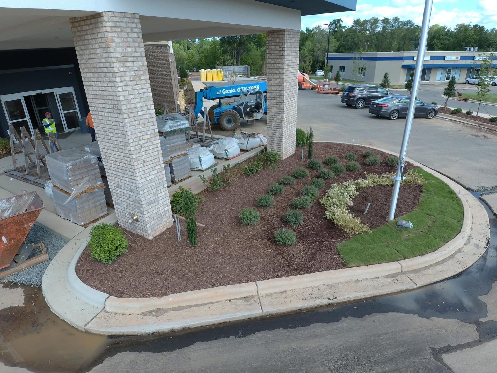 Landscaped entrance with brick columns, plants, and mulch. A blue lift and construction materials are in the background.