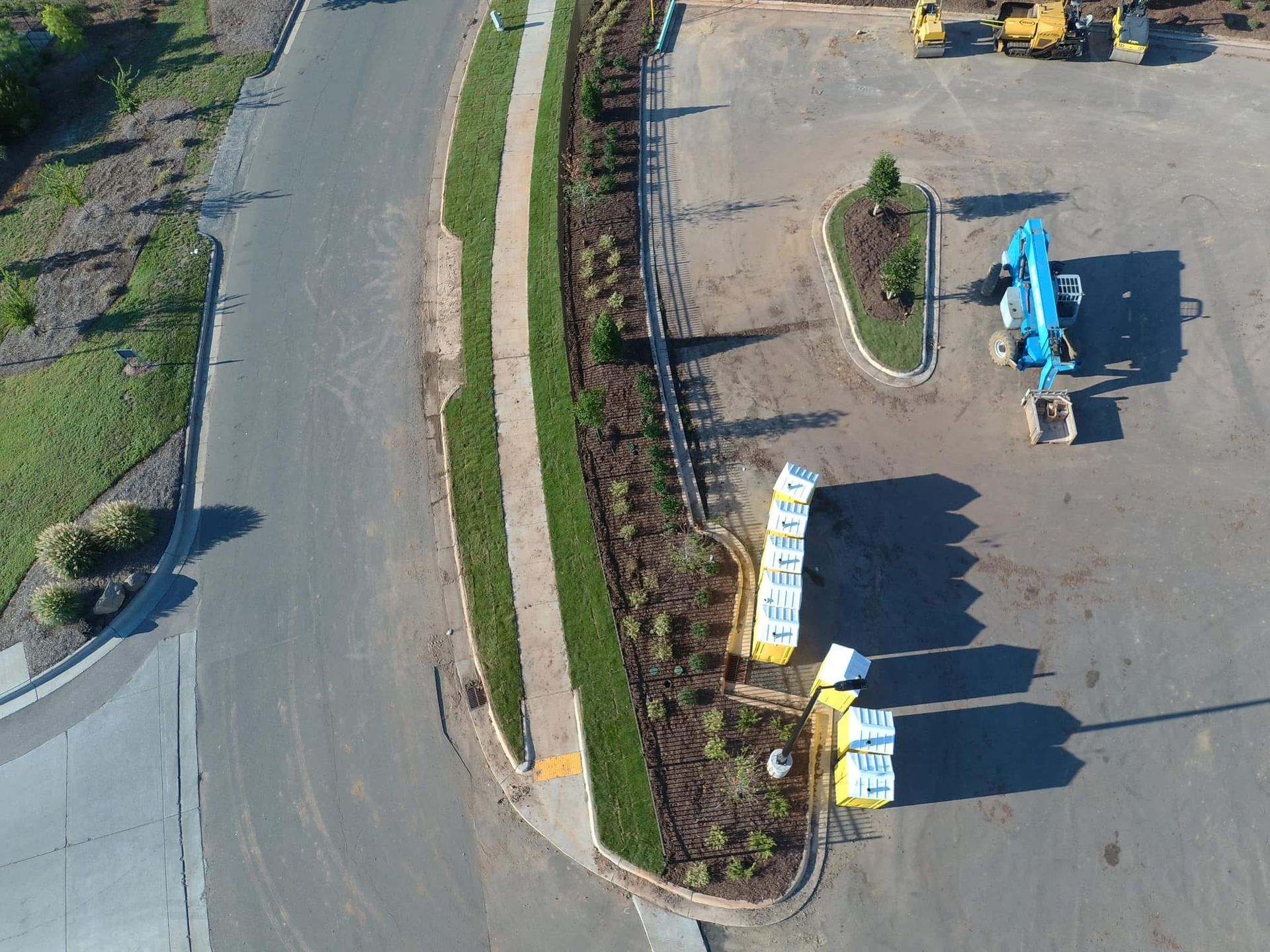 Aerial view: Asphalt road curving past a landscaped strip and a construction equipment parking area.