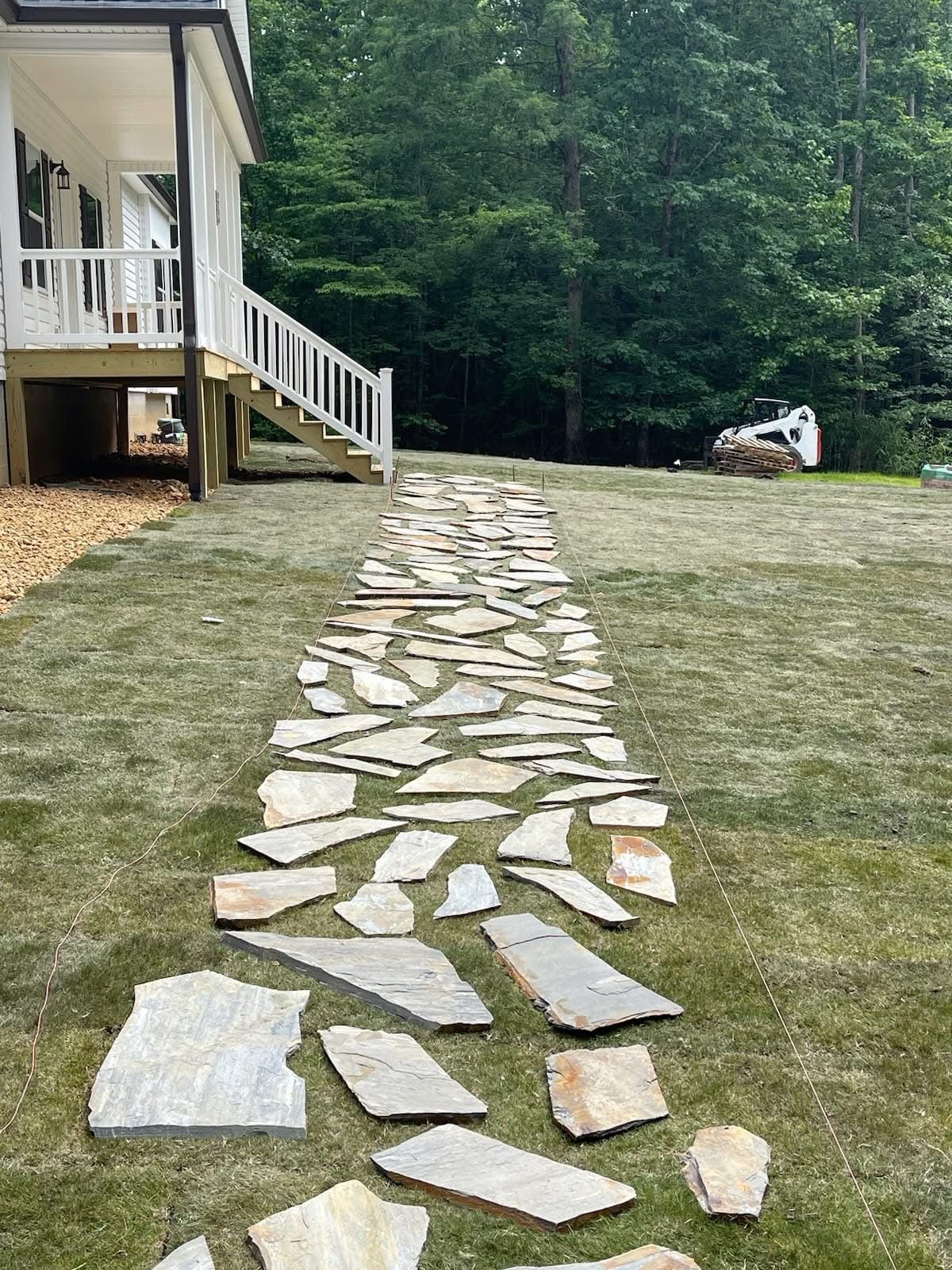 A stone pathway is being laid in the grass, leading from a house with a white porch toward a forest.