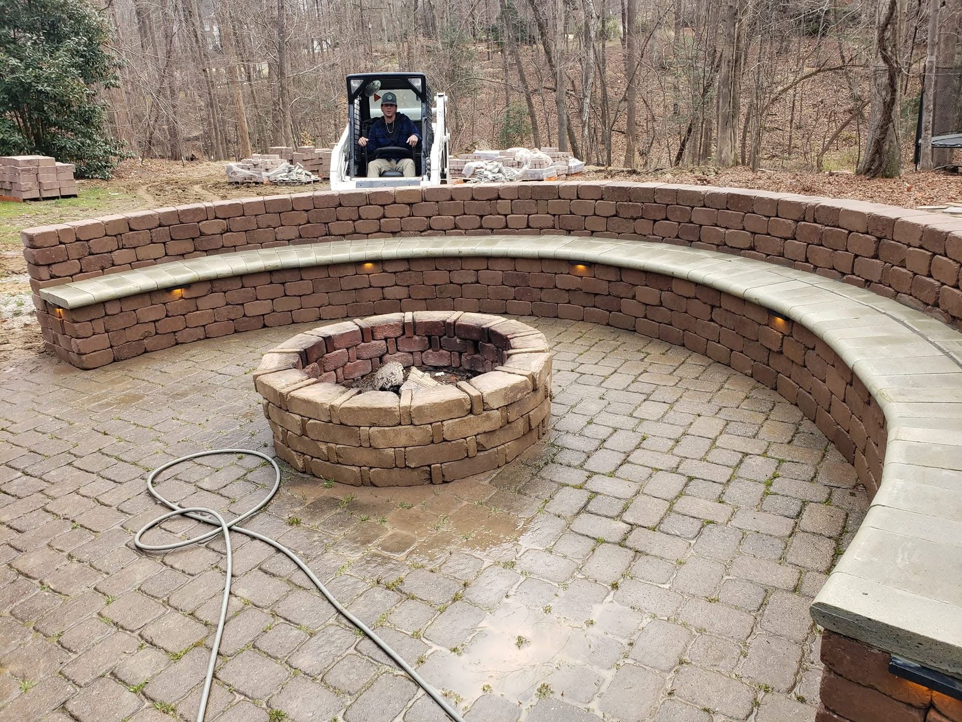 Curved brick seating around a fire pit on a paver patio; a person operates a small construction vehicle in the background.
