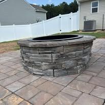 Stone fire pit on a brick patio in front of a house.