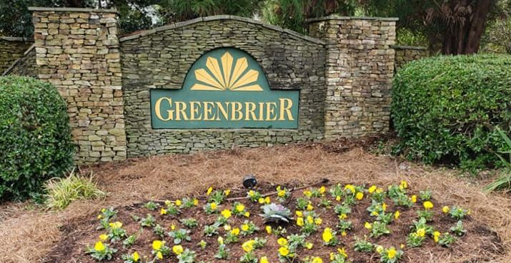 Sign for Greenbrier residential community. Green sign with gold design and text, set in stone entrance. Yellow flowers in foreground.