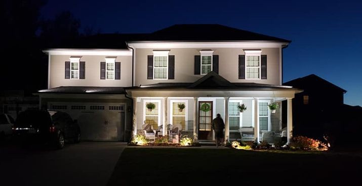 Two-story house illuminated at night. Beige siding, black shutters, white trim, porch with lights.