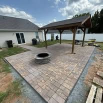 Patio with pavers, fire pit, and pergola next to a house with a white fence.