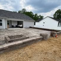 Backyard patio with stone fire pit and retaining walls. House in the background, overcast sky.