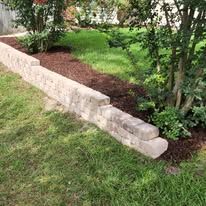 Stone retaining wall in a green yard, near a tree, with brown mulch and grass.