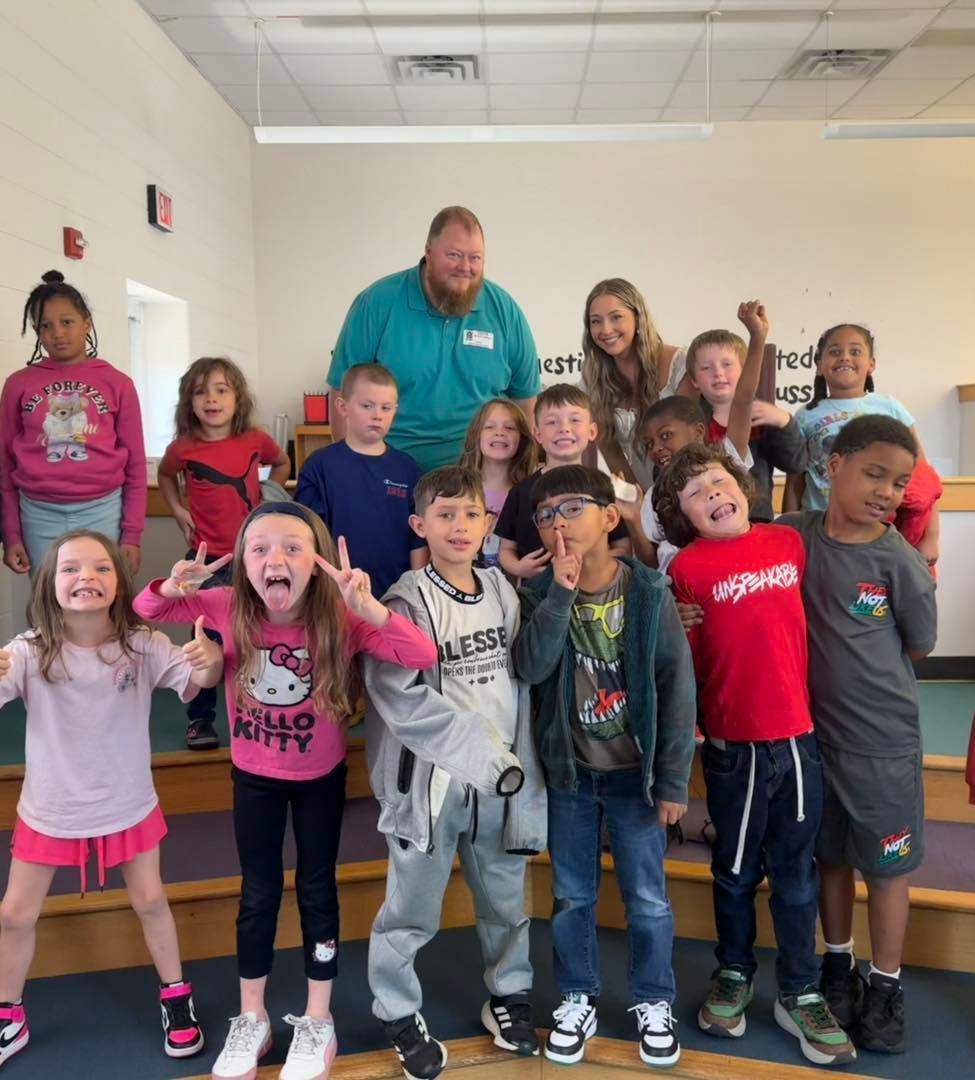 A group of children and two adults standing on tiered risers in a brightly lit room, smiling and making playful poses.