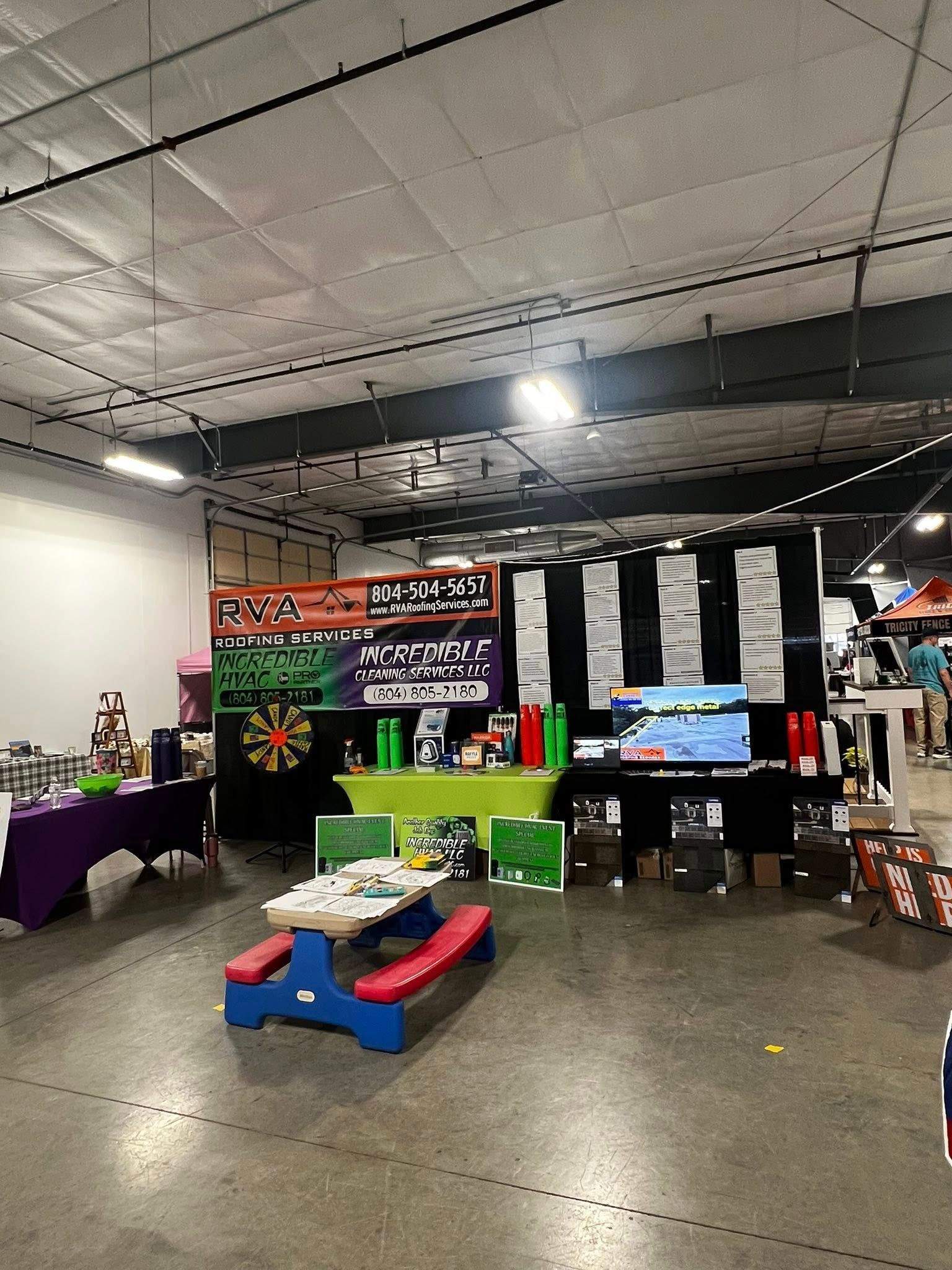 An indoor event space features a blue picnic table in the foreground, with a branded booth and display tables behind it.