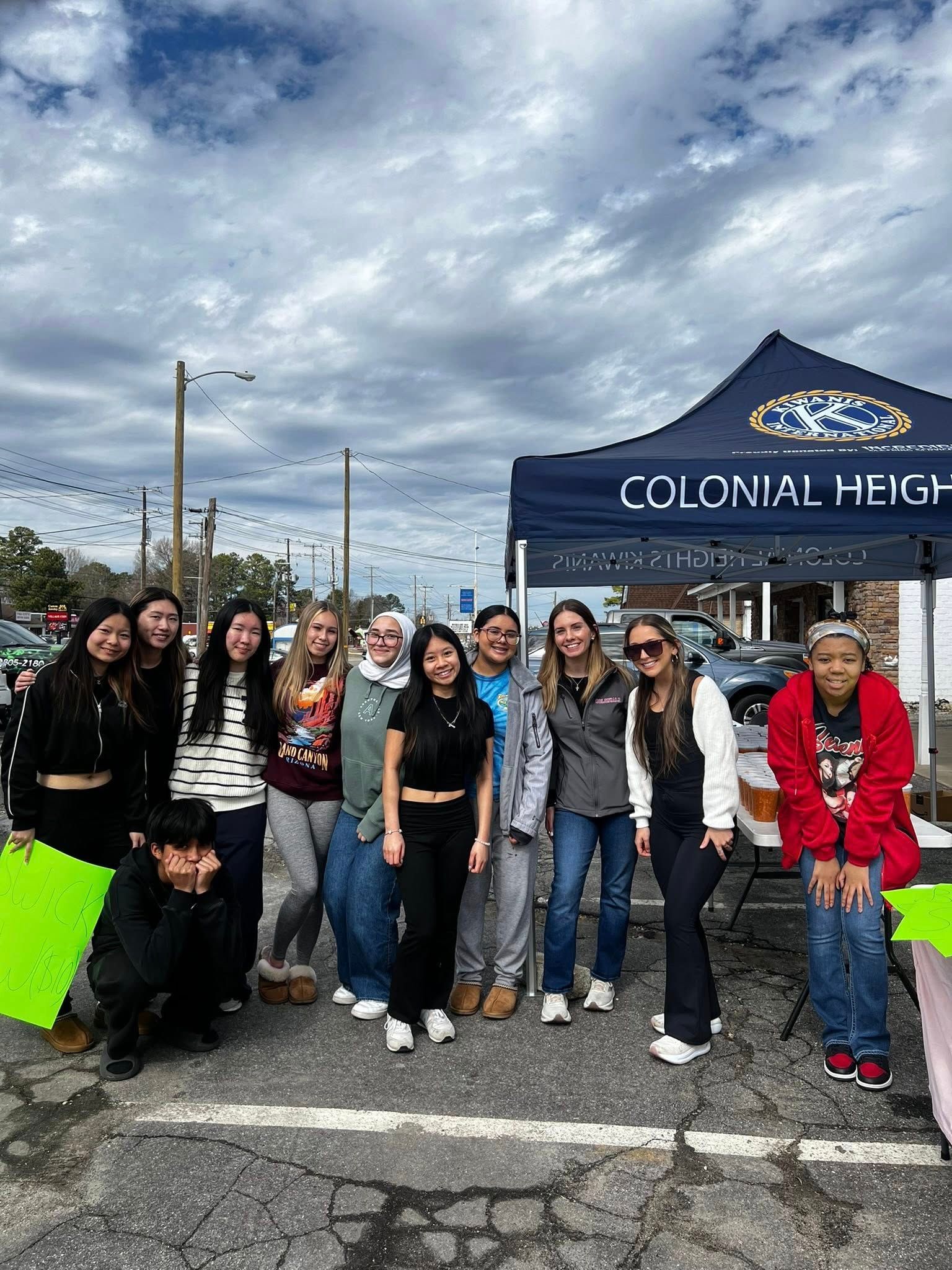 A group of people stand smiling together outdoors under a tent labeled Colonial Heights on a partly cloudy day.
