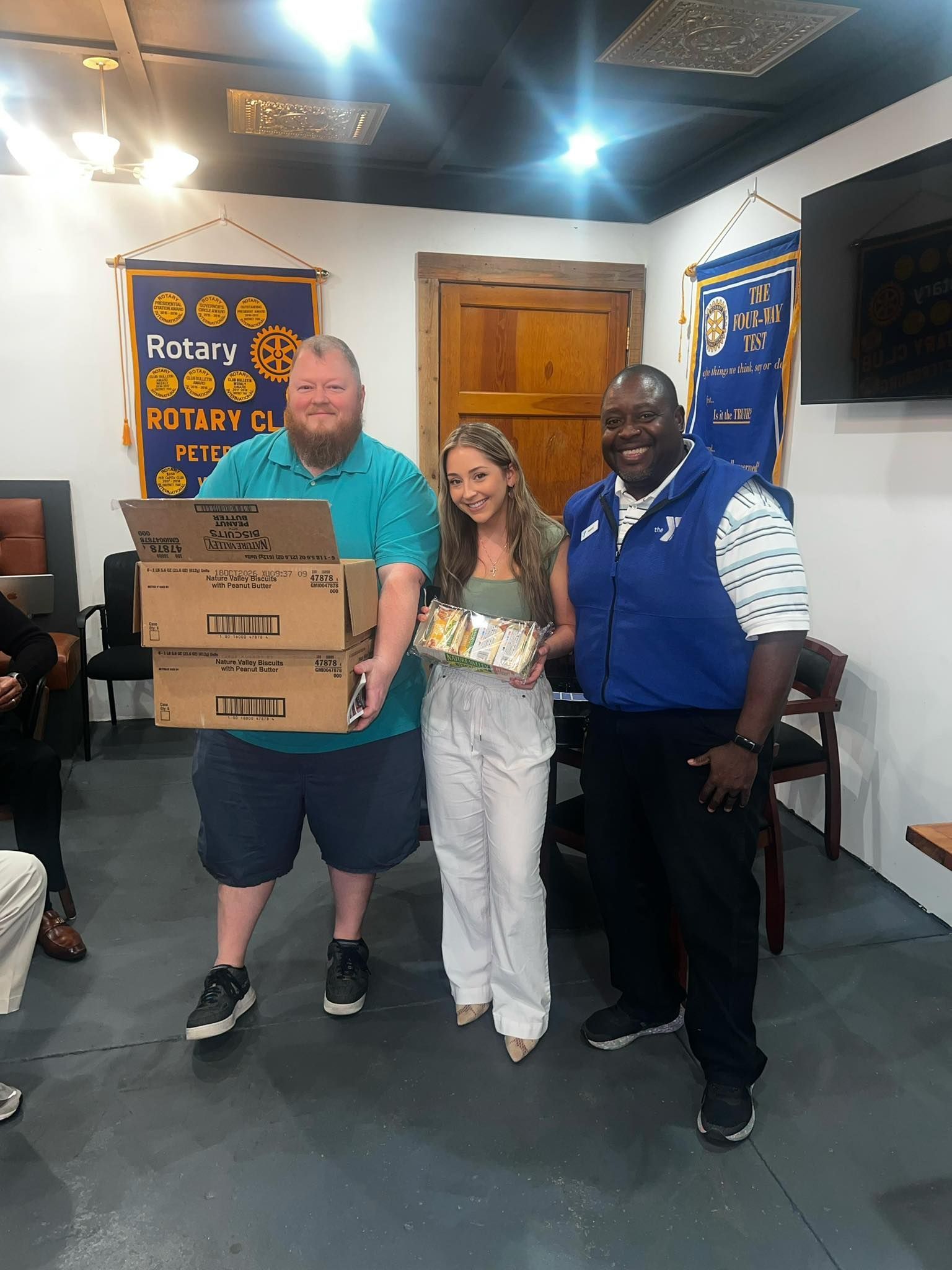 Three people stand in a Rotary Club meeting room, smiling while holding boxes and a tray of food.