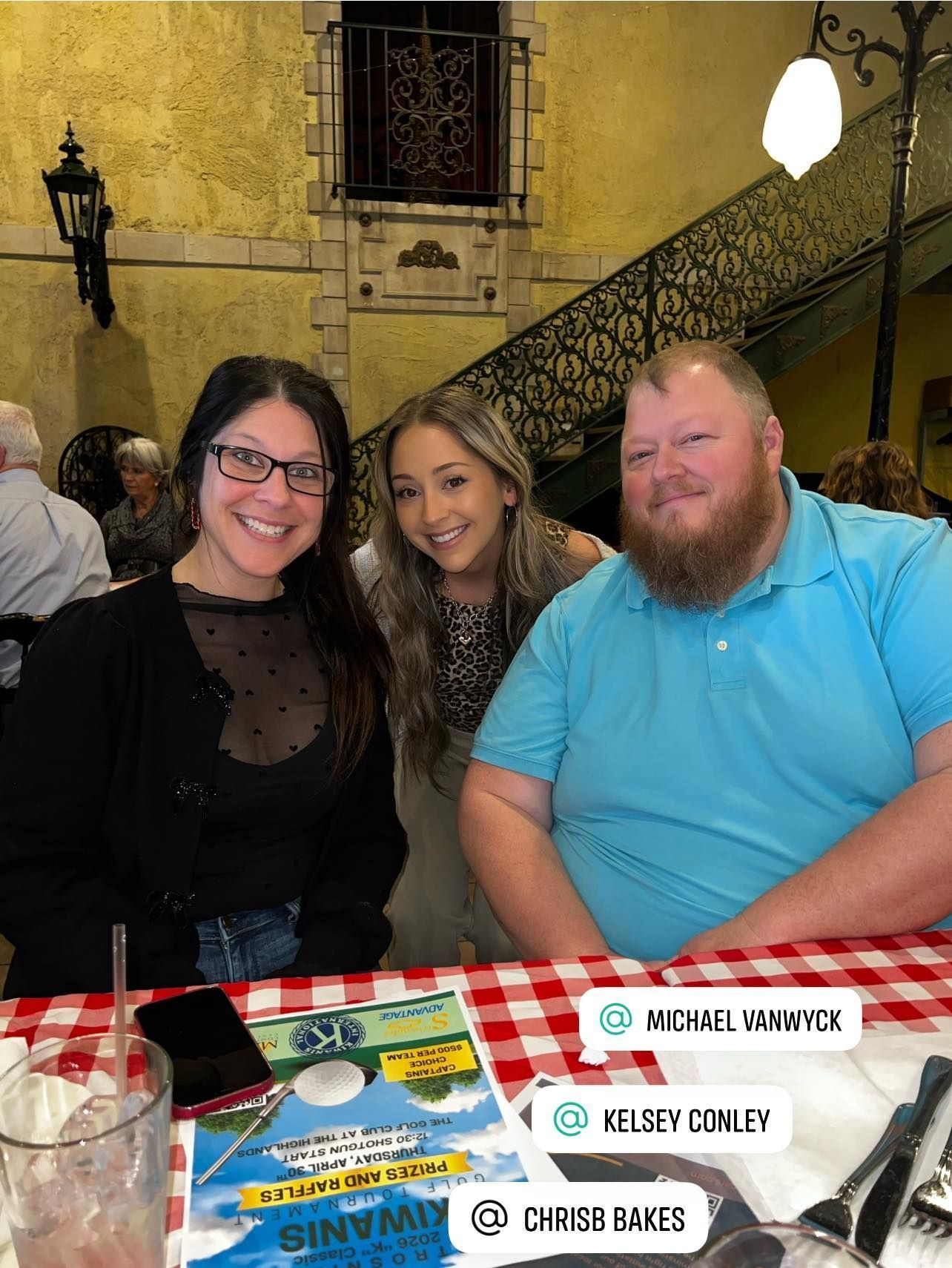 Three people smile at a table with a red-and-white checkered tablecloth at a Kiwanis event.