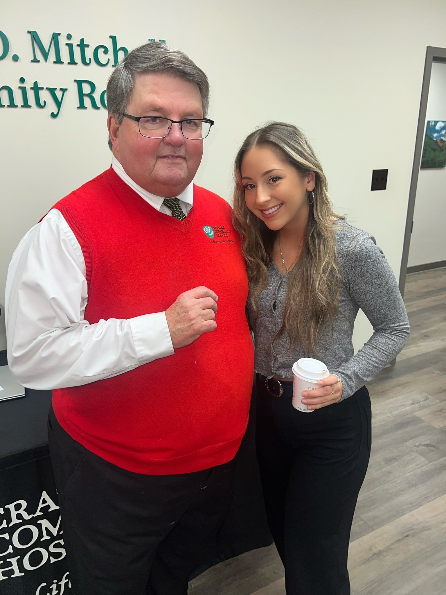 A man in a red sweater vest and a woman in a grey top stand together smiling in a room with a community room sign.
