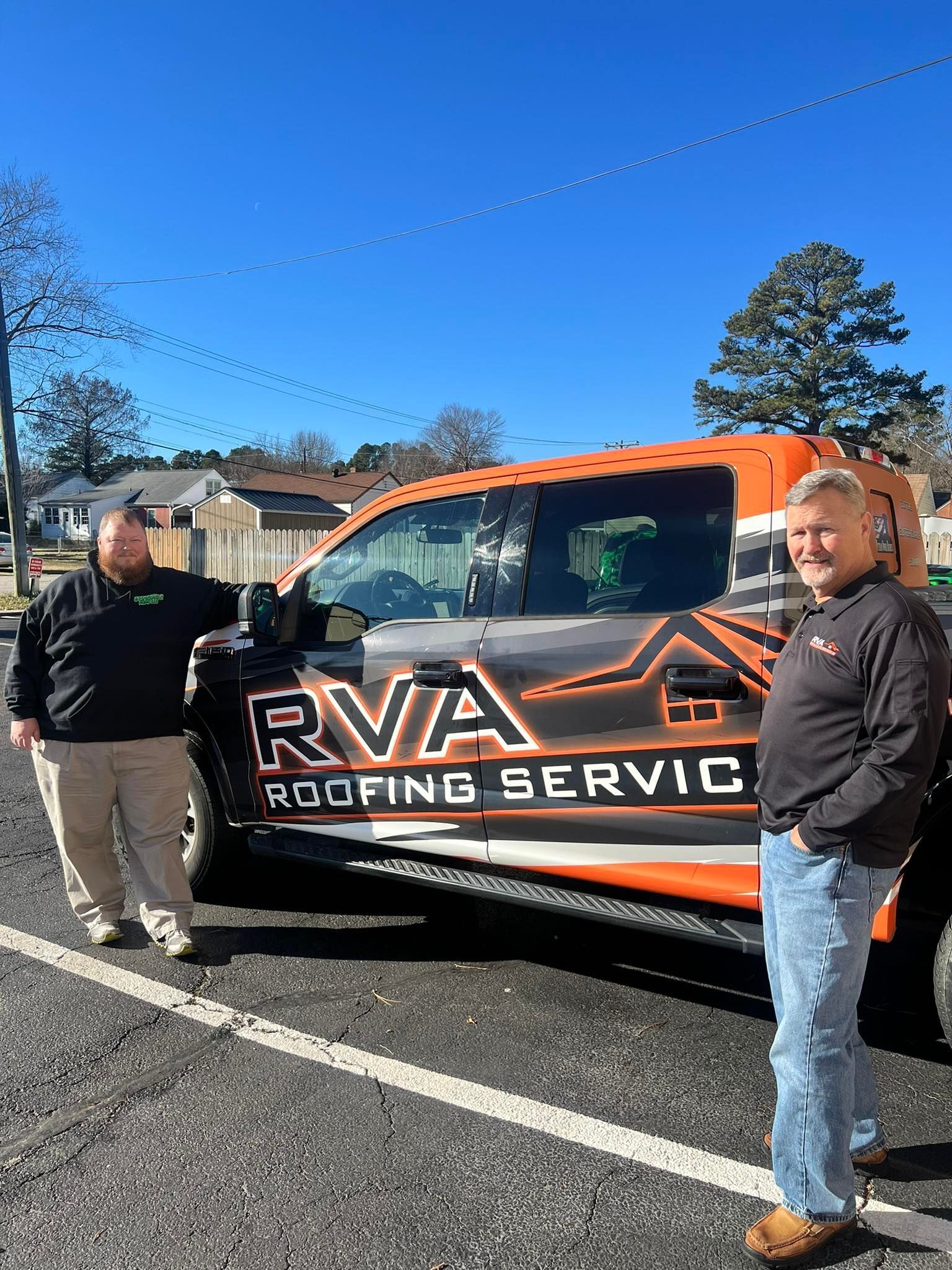 Two people stand next to an orange and black RVA Roofing Services pickup truck in an outdoor parking lot.