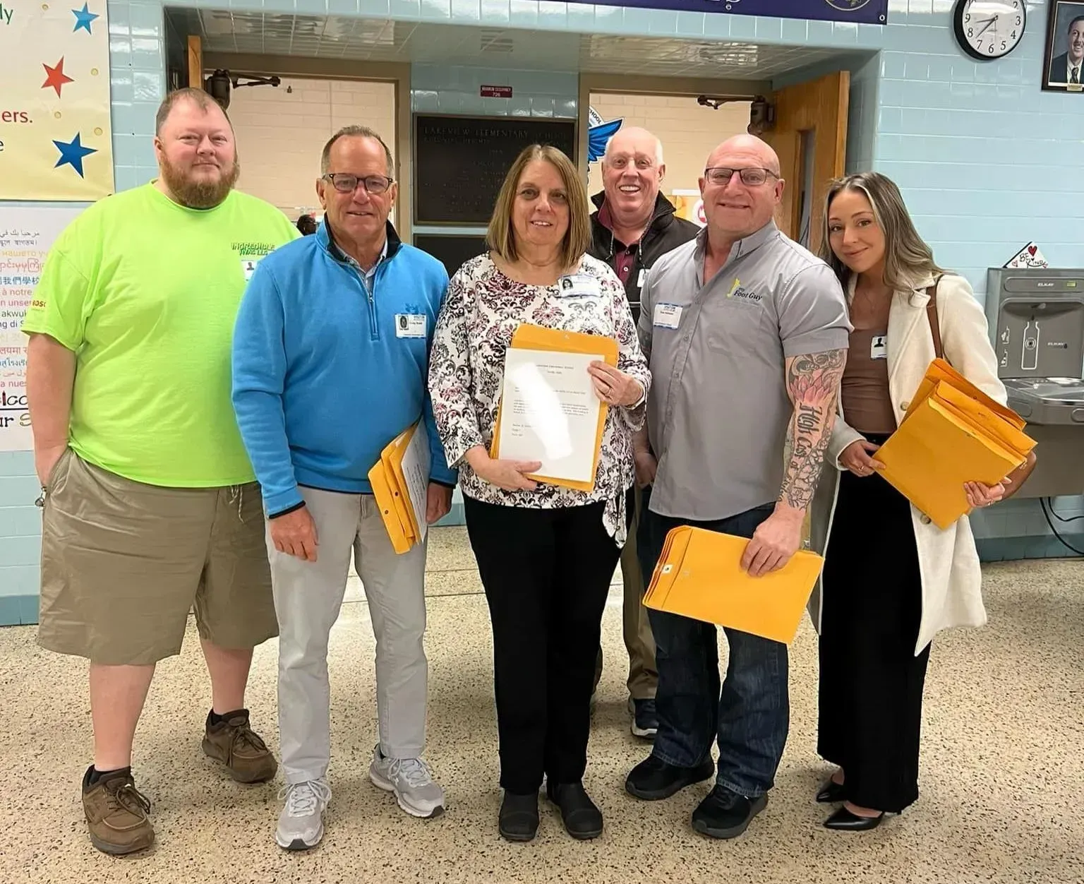 Group of people smiling, holding envelopes. Interior hallway setting, casual attire.