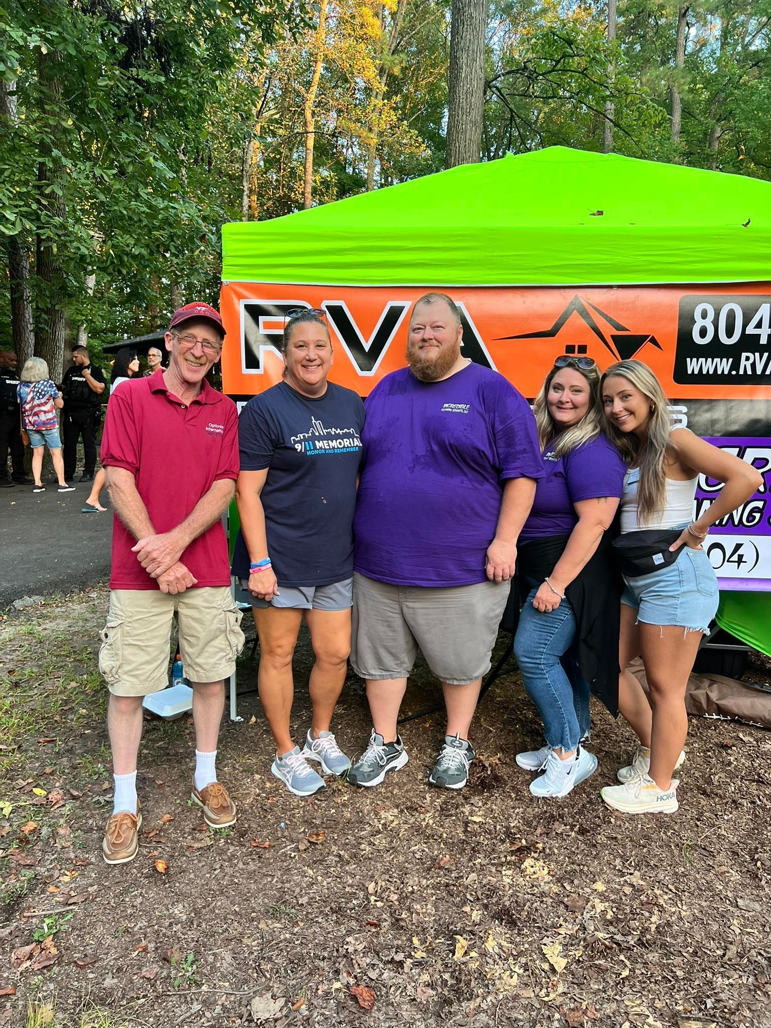 Group of six people posing near a green tent with a company logo. They wear casual clothes, with smiles. Forest in the background.