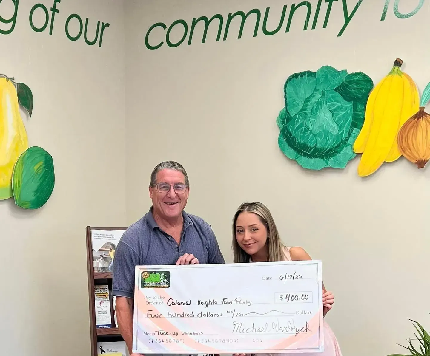 Man and young woman holding a large check, smiling in front of painted fruit on a wall.