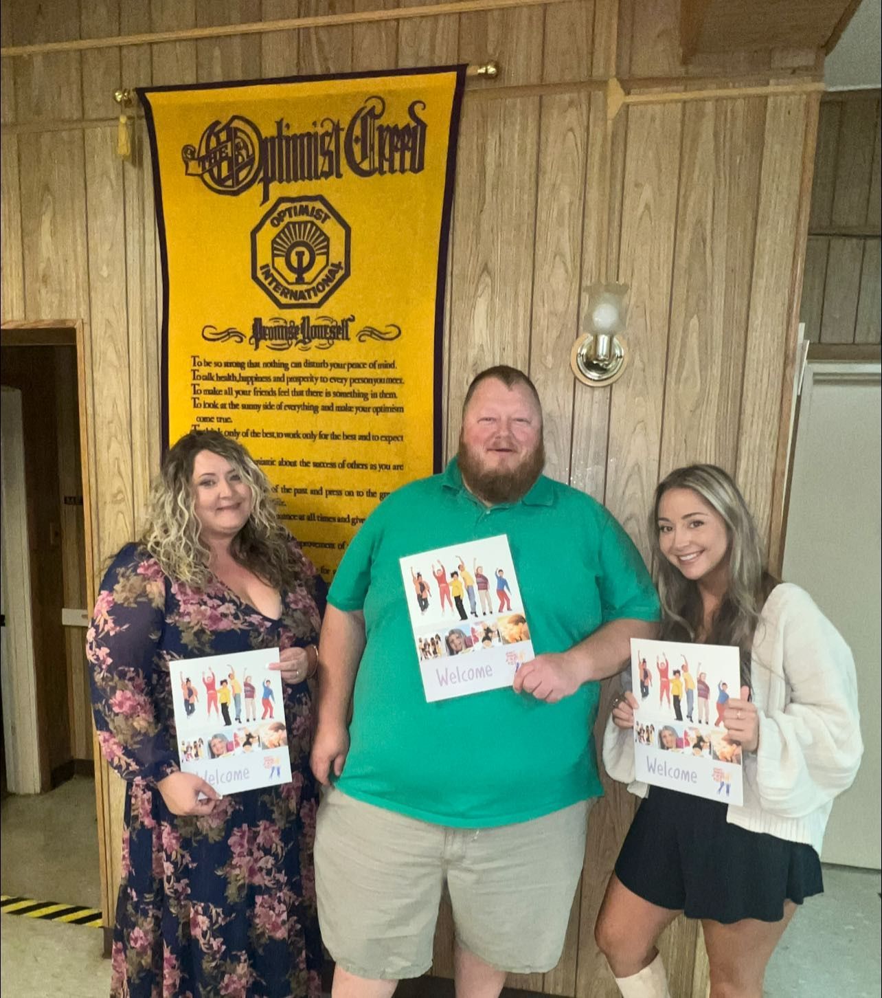 Three people holding posters, standing in front of an Optimist Creed banner. One is wearing a floral dress, the other a green shirt and khaki shorts, and the last, a white jacket and black skirt.