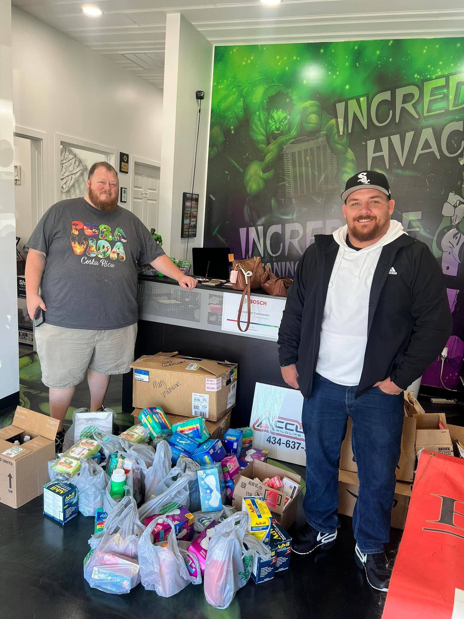 Two men stand with gifts in an office. One in a black jacket, the other in a t-shirt and shorts. Boxes and bags of presents are visible.