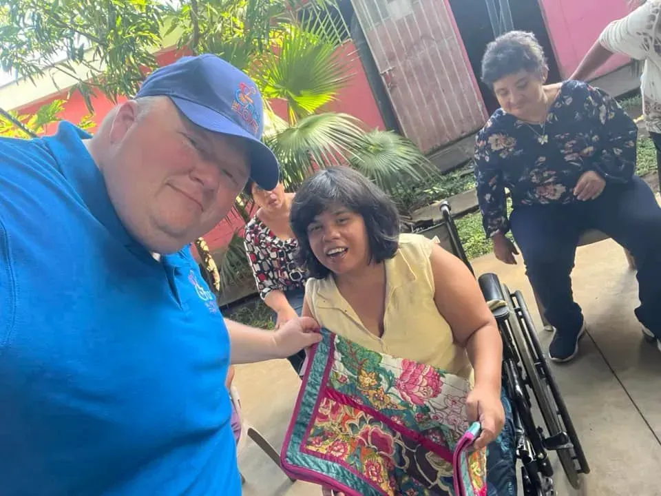 Man in blue shirt with group, including woman in wheelchair, holding quilt. Outdoor setting.