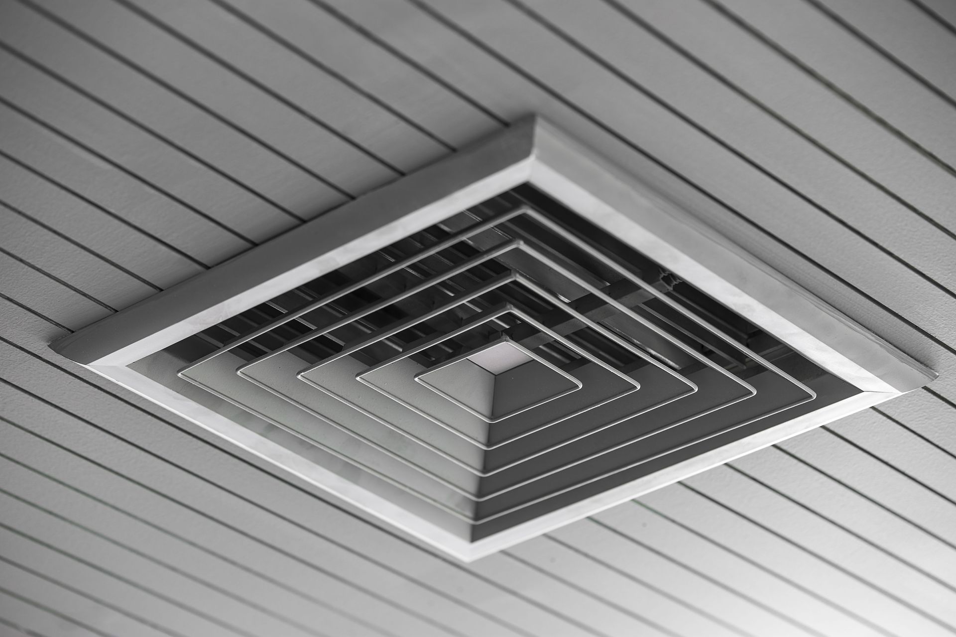 Square air vent recessed in a white, slatted ceiling.