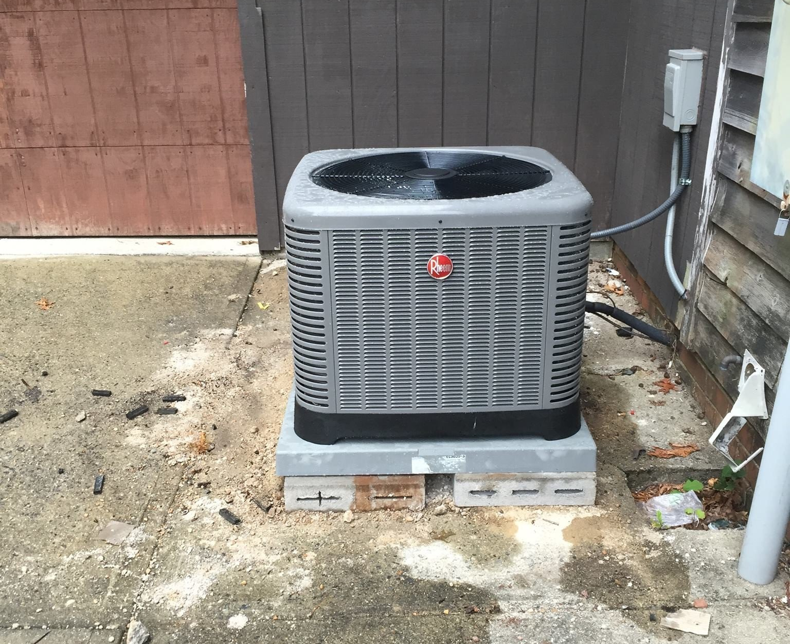 Gray Rheem air conditioner on a concrete base next to a wooden building, with water puddles on the ground.