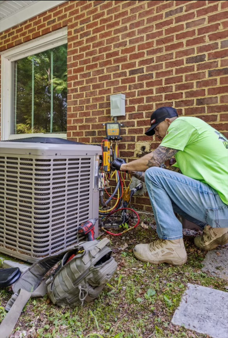 HVAC technician in a green shirt repairs an air conditioner unit against a brick wall outdoors.