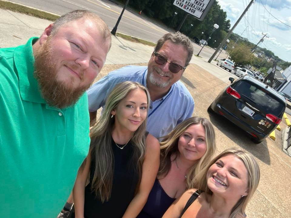 Five people posing for a selfie outside. Green shirt, blue shirt, black and navy tops. Sunny day with a car in the background.
