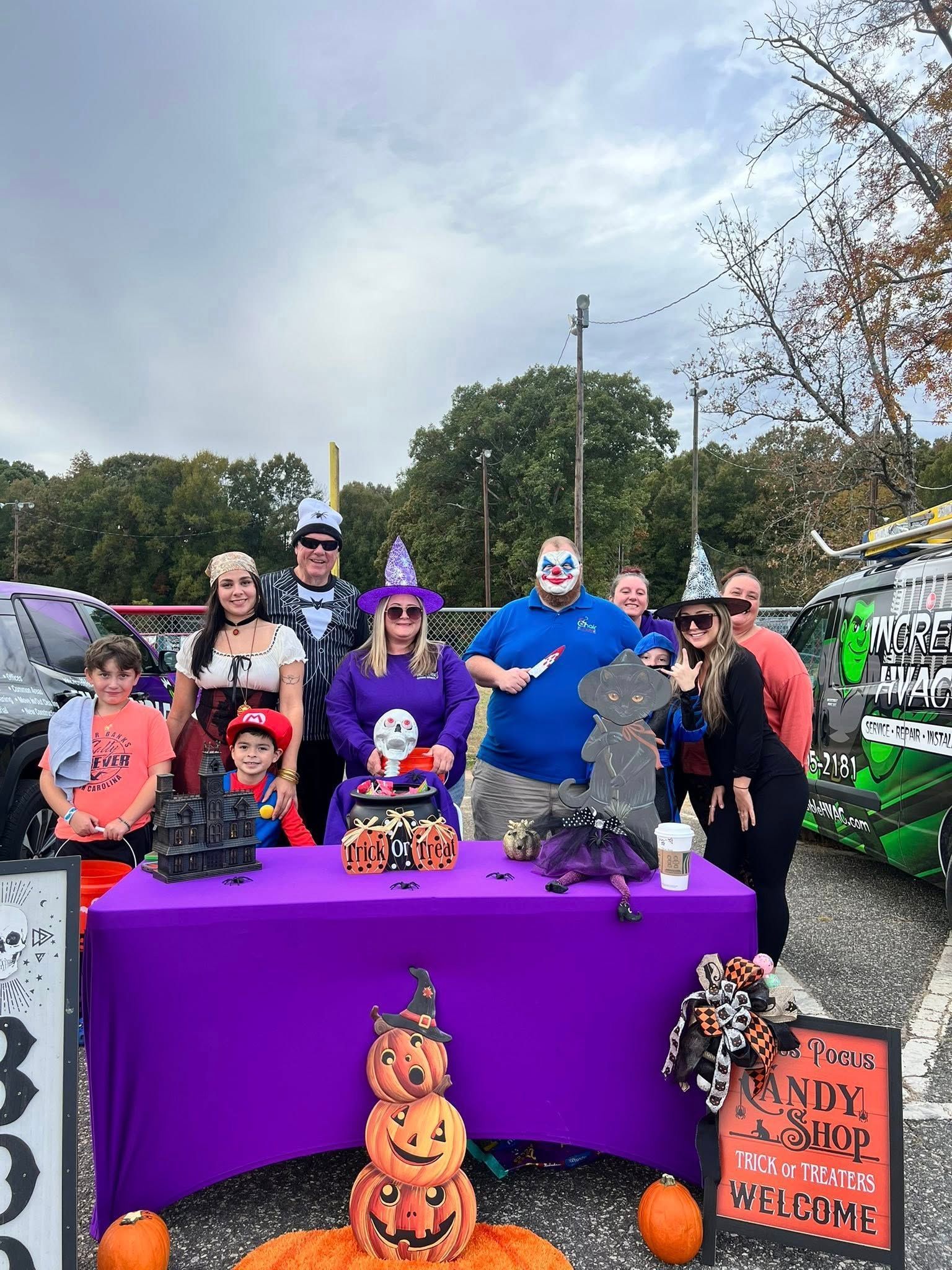 Group of people in Halloween costumes around a purple table. Pumpkins and decorations. Cloudy outdoor setting.